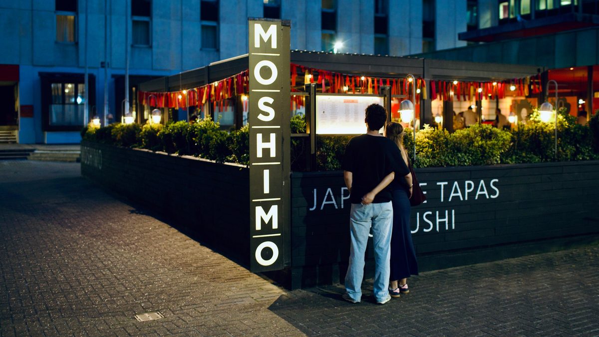 A couple reading the lit menu board outside Moshimo at night, the tall MOSHIMO sign beside them